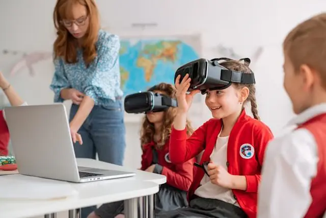 Children in a classroom trying VR headsets while a teacher works at a laptop.